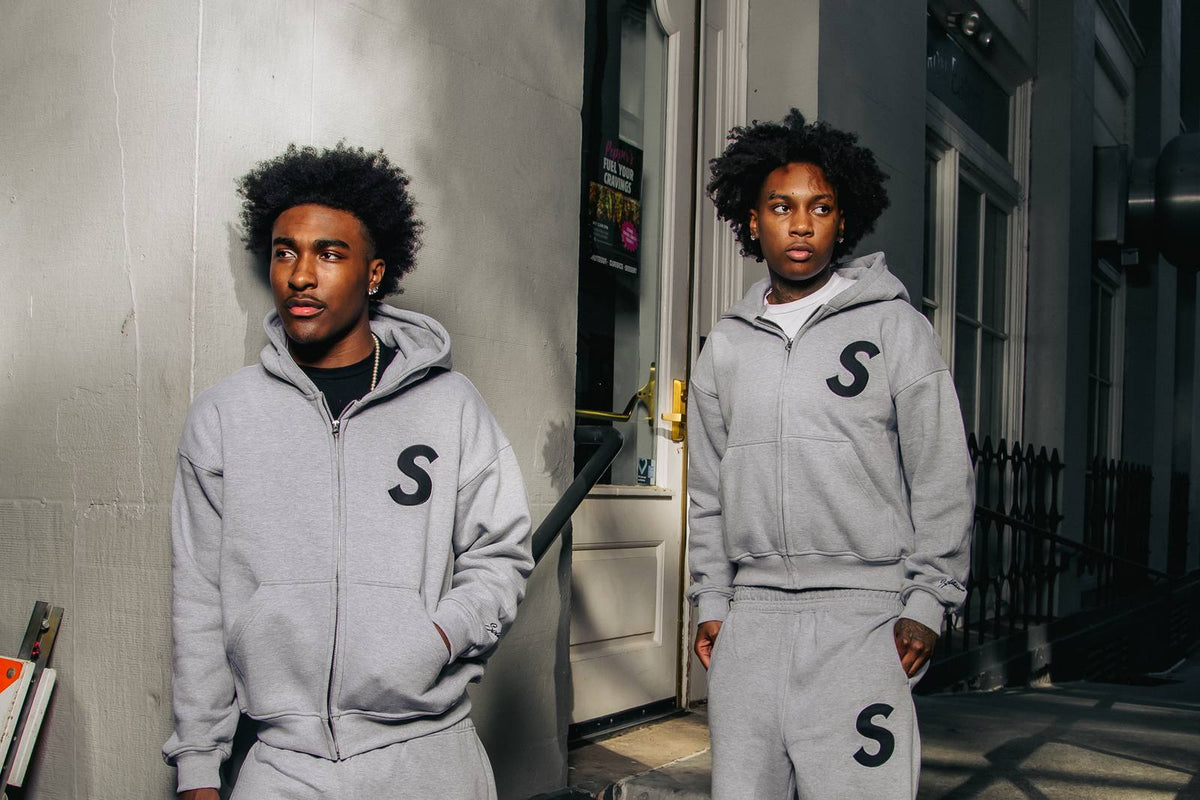 Two models wearing the grey Solitaire S hooded zip-up and matching shorts on a city street corner